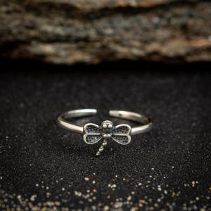 May include: A silver-colored ring featuring a dragonfly design. The dragonfly has detailed wings and a small body. The ring is adjustable and sits on a dark, textured surface.