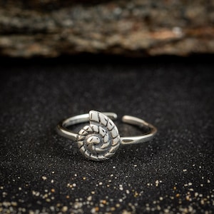 May include: A silver-colored ring featuring a detailed spiral seashell design. The ring is adjustable and sits on a dark, textured surface. The seashell design is the focal point, showcasing intricate patterns.