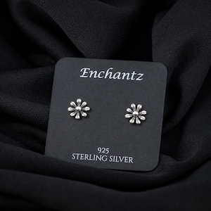 May include: A pair of silver daisy flower stud earrings on a black card with the text "Enchantz 925 Sterling Silver".