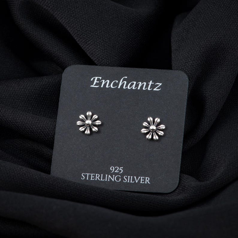 May include: A pair of silver daisy flower stud earrings on a black card with the text "Enchantz 925 Sterling Silver".