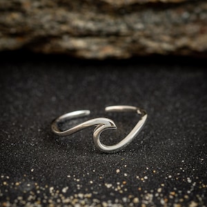 May include: A silver-colored ring shaped like a wave. The ring has a smooth, polished finish and a minimalist design. The wave crest is the focal point, creating a nautical or beach-themed accessory.