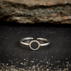 May include: A silver-colored adjustable ring with a circular design. The ring is set against a dark, textured background, with a blurred wooden element in the background. The ring's design is simple and modern.