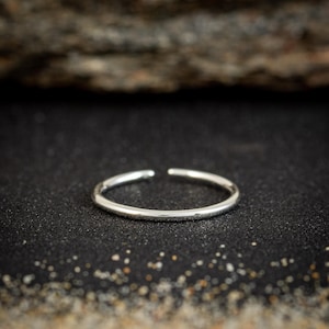 May include: A minimalist silver ring with a slight opening. The ring is set against a dark, textured surface, with a blurred background. The ring appears to be made of a shiny metal.