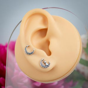 K&ouml;nnte beinhalten: Ein Paar silberne Ohrringe mit einem Halbmond-Design. Ein Ohrring hat ein schlichtes Mond-Design, w&auml;hrend der andere eine Katze auf dem Mond hat.