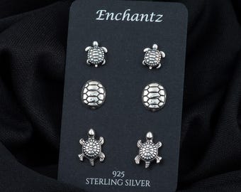 May include: A set of six sterling silver turtle earrings. The earrings feature three different designs: a turtle with its shell facing up, a turtle with its shell facing down, and a turtle with its shell facing sideways. The earrings are displayed on a black card with the word "Enchantz" and the text "925 Sterling Silver".