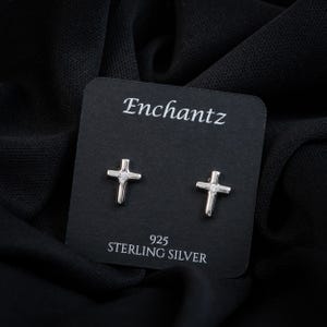 May include: A pair of sterling silver cross stud earrings with a small clear gemstone in the center of each cross. The earrings are displayed on a black card with the brand name "Enchantz" and the text "925 STERLING SILVER".