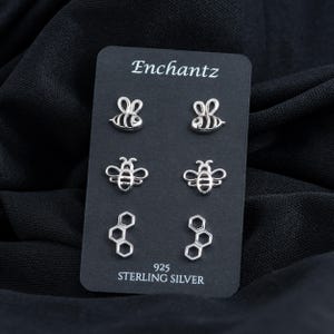 May include: A black card with the word "Enchantz" printed on it, displaying six pairs of silver-colored stud earrings. The earrings are shaped like bees and honeycomb. The card also has the text "925 STERLING SILVER".