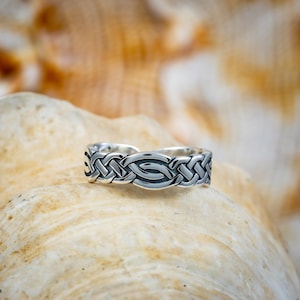 May include: A silver-colored ring with a Celtic knot design. The ring features an interwoven pattern with a central oval shape. The ring is displayed on a light-colored, textured surface, possibly a seashell.