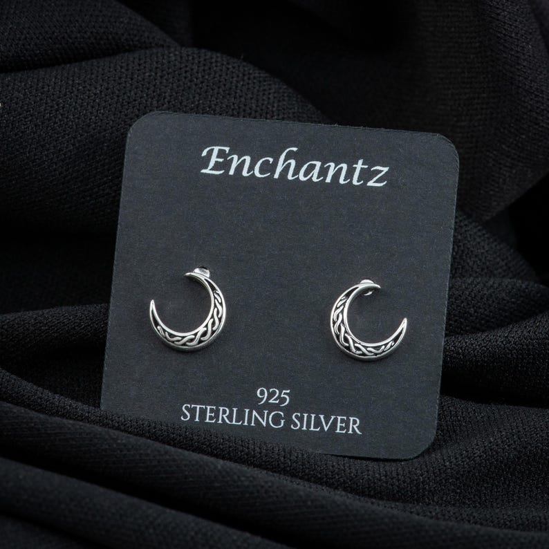 K&ouml;nnte beinhalten: Ein Paar Ohrringe aus Sterlingsilber mit einem Halbmond-Design mit komplizierten Knotenarbeiten. Die Ohrringe werden auf einer schwarzen Karte mit dem Wort "Enchantz" und "925 Sterling Silver" darauf pr&auml;sentiert.