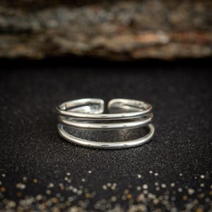 May include: A silver-colored ring with three bands. The ring is adjustable and sits on a dark, textured surface. The background features a blurred wooden texture.