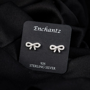 May include: A pair of sterling silver bow-shaped stud earrings on a black card. The card reads "Enchantz" at the top and "925 STERLING SILVER" at the bottom. The earrings are small and feature a delicate design.
