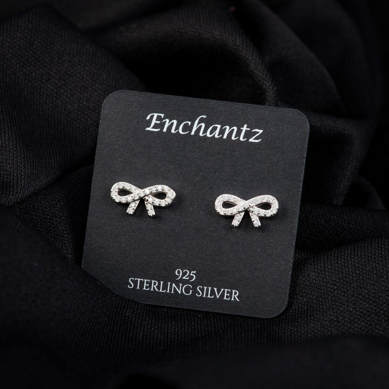 May include: A pair of sterling silver bow-shaped stud earrings on a black card. The card reads "Enchantz" at the top and "925 STERLING SILVER" at the bottom. The earrings are small and feature a delicate design.