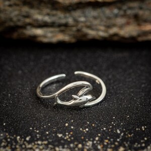 May include: A silver-toned open ring featuring a polished dolphin motif integrated into the band. The design shows the dolphin leaping through a curved wave shape, creating a continuous, fluid silhouette that wraps around the finger.