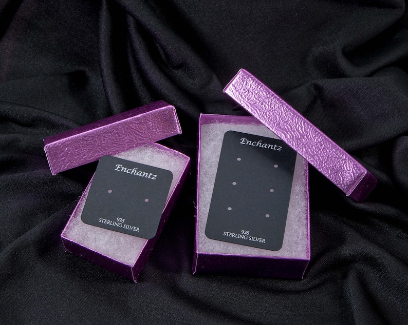 May include: Two purple earring boxes with black labels that say "Enchantz 925 Sterling Silver". The boxes are open and show the earring posts inside.
