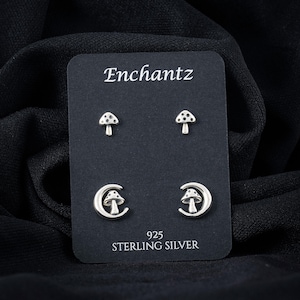 May include: A set of silver-colored mushroom earrings on a black card. The card reads "Enchantz" at the top and "925 STERLING SILVER" at the bottom. The earrings include two mushroom studs and two crescent moon designs with mushrooms.