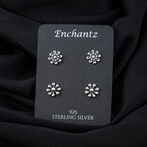 May include: A black card with the text "Enchantz" and "925 Sterling Silver" printed on it. Four silver flower-shaped earrings are displayed on the card.