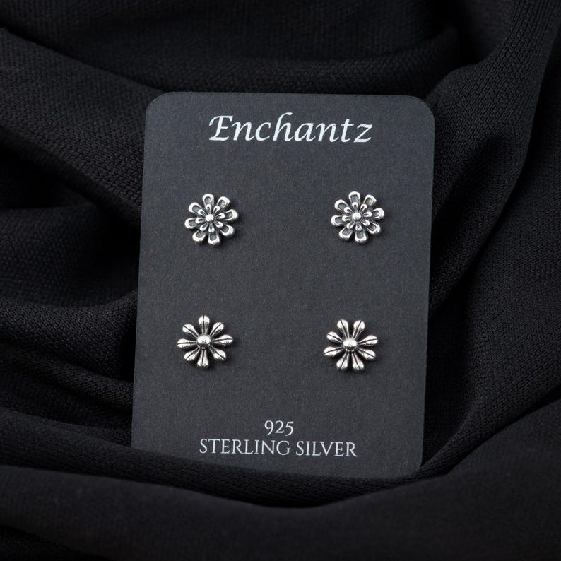 May include: A black card with the text "Enchantz" and "925 Sterling Silver" printed on it. Four silver flower-shaped earrings are displayed on the card.