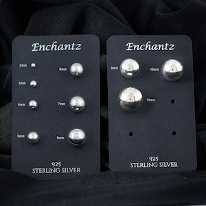 May include: Two black cards displaying sterling silver ball stud earrings. The cards are labeled "Enchantz" and "925 Sterling Silver." The earrings range in size from 2mm to 11mm. The earrings are shiny and reflective.