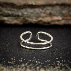 May include: A silver-colored double-band ring. The ring has a minimalist design with two parallel bands that curve around the finger. The ring is set against a dark, textured background.