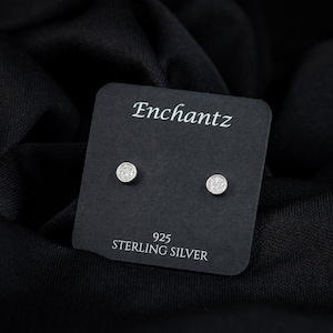 May include: A pair of round, silver-colored stud earrings with a sparkling, textured surface. The earrings are displayed on a black card with the brand name "Enchantz" and the text "925 STERLING SILVER". The background is a black fabric.