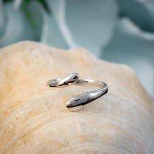 May include: A silver-colored ring with a fluid, open design. The ring features two teardrop-shaped ends that curve around to meet. The ring is displayed on a light-colored, textured surface, with a soft blue background.