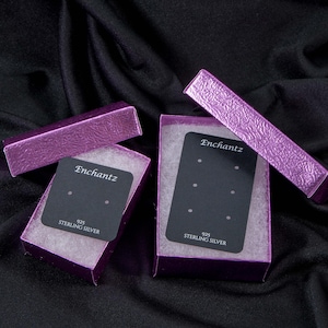 May include: Two open purple jewelry boxes with black earring display cards. The boxes are lined with white material. The cards have the word "Enchantz" printed on them, along with "925 Sterling Silver". The boxes are on a black background.
