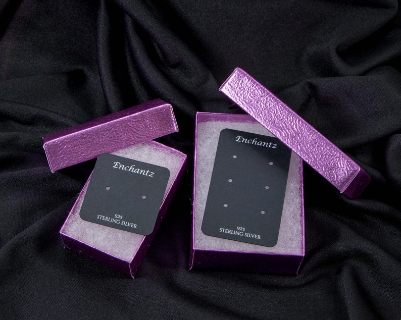 May include: Two open purple jewelry boxes with black earring display cards. The boxes are lined with white material. The cards have the word "Enchantz" printed on them, along with "925 Sterling Silver". The boxes are on a black background.