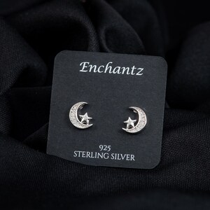 May include: A pair of sterling silver stud earrings featuring a crescent moon and star design. The earrings are displayed on a black card with the brand name "Enchantz" and "925 Sterling Silver" printed on it. The background is a black fabric.