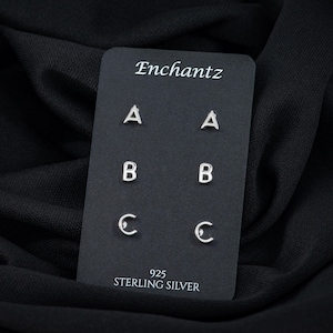 May include: Silver letter stud earrings displayed on a black card. The card reads "Enchantz" at the top and features the letters A, B, and C. The bottom of the card reads "925 STERLING SILVER".