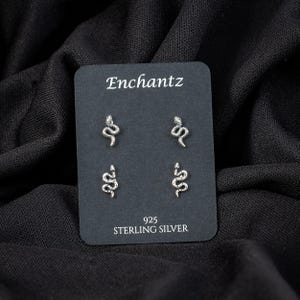 May include: Silver snake stud earrings on a black display card. The card reads "Enchantz" at the top and "925 STERLING SILVER" at the bottom. The earrings are small, detailed, and feature a coiled snake design.