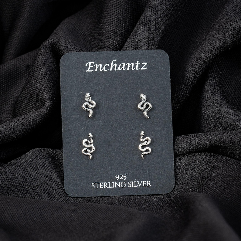May include: Silver snake stud earrings on a black display card. The card reads "Enchantz" at the top and "925 STERLING SILVER" at the bottom. The earrings are small, detailed, and feature a coiled snake design.
