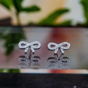 May include: A pair of silver bow-shaped stud earrings. Each earring is encrusted with small, clear gemstones, creating a sparkling effect. The earrings are displayed on a reflective surface, with a blurred green and white background.