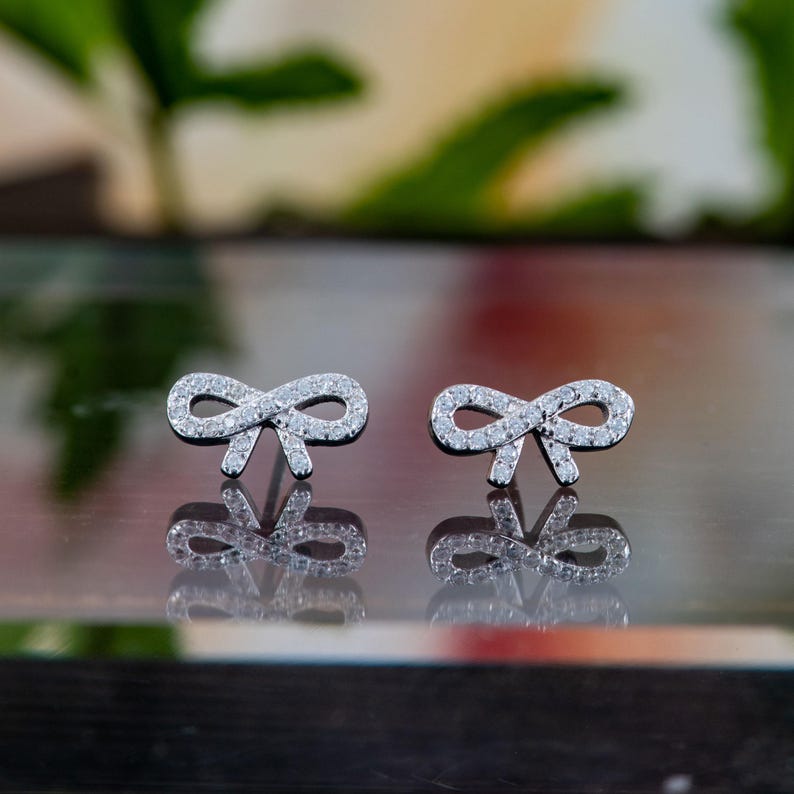 May include: A pair of silver bow-shaped stud earrings. Each earring is encrusted with small, clear gemstones, creating a sparkling effect. The earrings are displayed on a reflective surface, with a blurred green and white background.