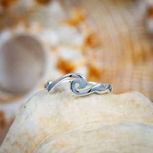 May include: A silver ring with a wave design. The ring is set against a blurred background of a seashell. The wave design is a continuous line that forms a curling wave shape. The ring is a simple, elegant piece of jewelry.