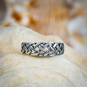 May include: A silver ring with a woven design. The ring features an intricate pattern of interwoven metal bands, creating a textured and detailed appearance. The ring is displayed on a light-colored surface, highlighting its metallic sheen.