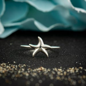 May include: A silver starfish ring. The ring features a detailed starfish design, with five arms, set on a thin band. The ring is displayed on a black surface with a blurred blue background and a scattering of sand-like particles.