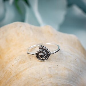 May include: A silver-colored ring with a spiral seashell design. The ring is set on a light tan surface, with a blurred blue and green background. The seashell design is detailed and textured.