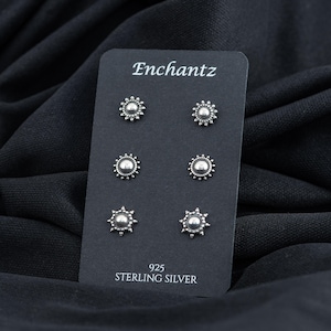 May include: A set of six silver stud earrings with a sunburst design. The earrings are displayed on a black card with the word "Enchantz" and "925 Sterling Silver" printed on it.