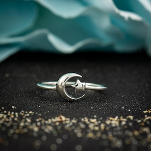 May include: A silver ring featuring a crescent moon and star design. The ring is set against a dark background with a blurred blue backdrop, creating a celestial theme. The ring's design is simple yet elegant.
