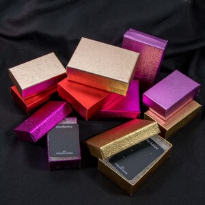 May include: A collection of small, rectangular gift boxes in various colors, including gold, red, pink, and purple. Some boxes are open, revealing black display cards. The boxes have a textured, metallic finish, and the text "Enchantz" is visible on one of the cards.