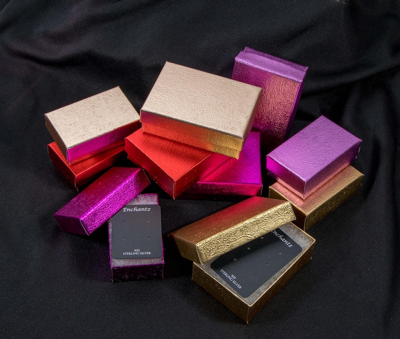 May include: A collection of small, rectangular gift boxes in various colors, including gold, red, pink, and purple. Some boxes are open, revealing black display cards. The boxes have a textured, metallic finish, and the text "Enchantz" is visible on one of the cards.
