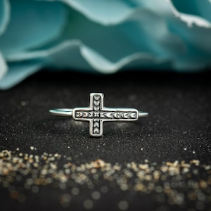 May include: A silver-toned ring featuring a horizontal cross centerpiece. The cross is decorated with a repeating pattern of small, leaf-like indentations along its entire surface, creating a textured appearance on the metal band.