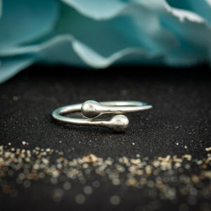 May include: A silver-colored ring with a minimalist design. The ring features two teardrop-shaped ends that overlap. The ring is set against a black surface with a blurred blue background.