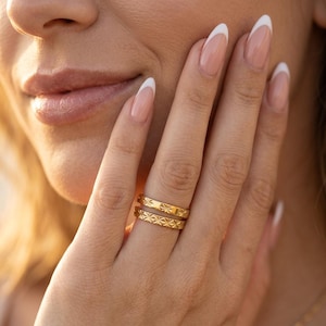 Puede incluir: Primer plano de una mano que lleva dos anillos de oro. Los anillos tienen un patrón de estrellas repetido. Las uñas están pintadas con una manicura francesa. El tono de la piel es claro y el fondo está borroso.