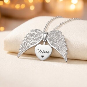 May include: A silver-colored necklace with a heart-shaped pendant and angel wings. The heart pendant is engraved with the name "Marie" and a small heart. The wings are embellished with small, clear crystals.
