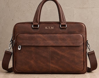May include: A brown leather briefcase featuring two top handles, a detachable shoulder strap with silver-toned metal hardware, and a front zippered pocket. The center of the bag is embossed with the letters K.T.M in a simple, clean font.