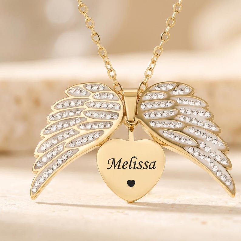May include: A gold-colored necklace with a heart-shaped pendant and angel wings. The pendant is engraved with the name "Melissa" and a small black heart. The wings are adorned with clear crystals.