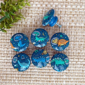 May include: Seven blue and white dinosaur themed cabinet knobs with a space theme. The knobs feature different dinosaurs, stars, planets, and moons.