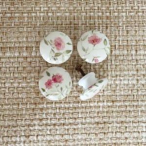 May include: Four round ceramic cabinet knobs featuring a white base with a hand-painted floral pattern of pink roses and green leaves. Three knobs are shown from the front, while one knob is displayed from the side, revealing a metal screw.