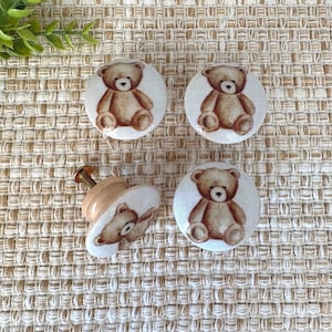 May include: Four decorative cabinet knobs and drawer pulls with a teddy bear design. The knobs are round with a light beige background and a brown teddy bear illustration. One knob is attached to a wooden base with a screw.